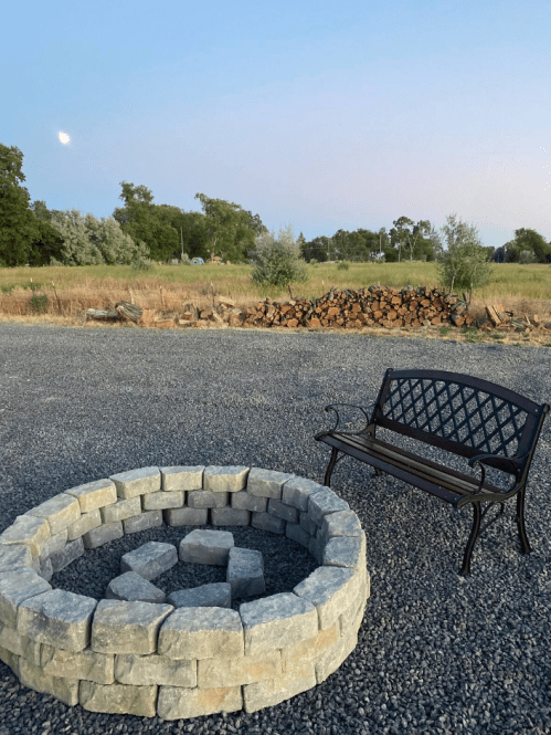 A circular stone fire pit beside a black metal bench, set in a gravel area with trees and a moonlit sky in the background.